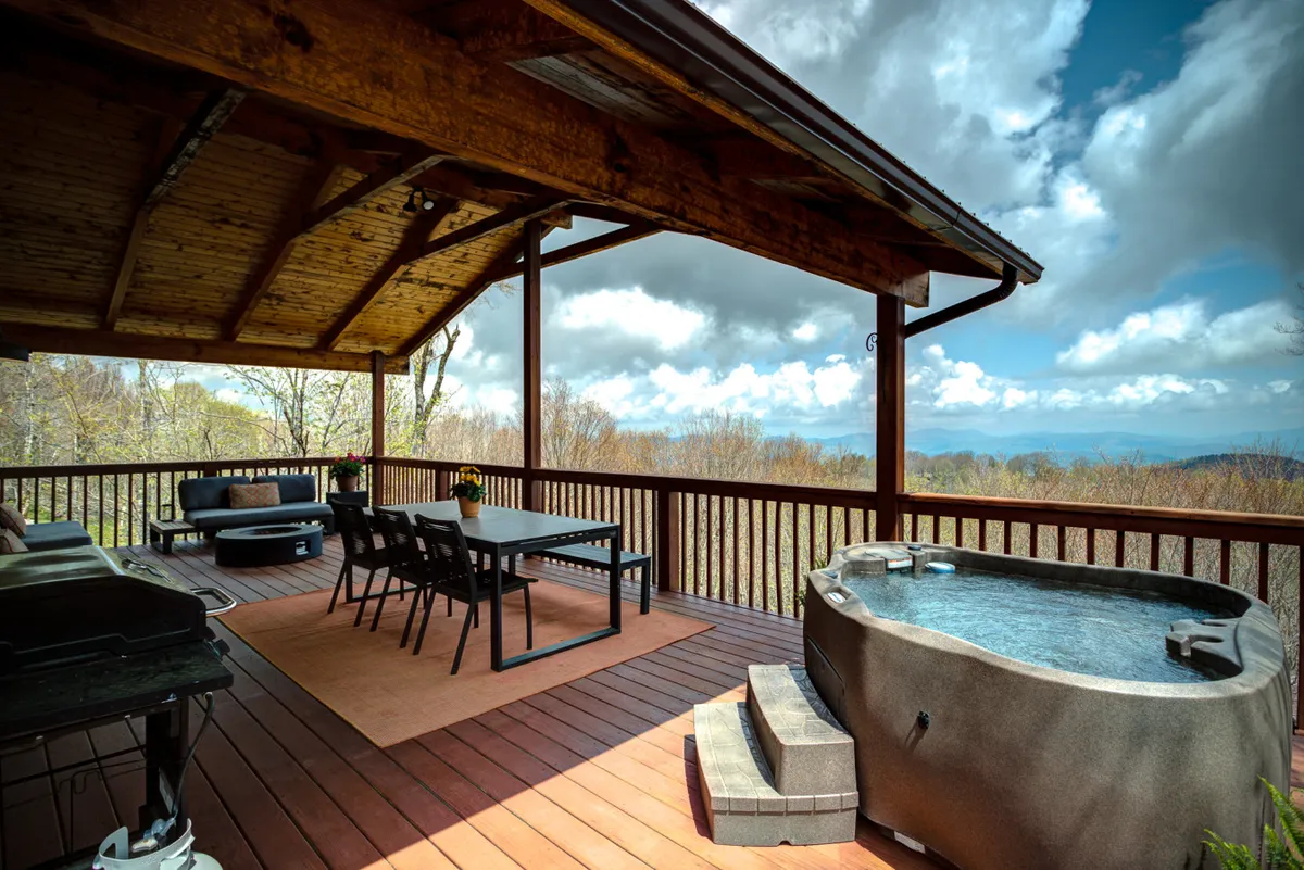 The image shows an inviting chalet deck at sunset, featuring a bubbling hot tub with panoramic mountain views, a spacious outdoor dining table, cozy seating, and a grill—perfect for relaxing, dining, and enjoying the serene surroundings.