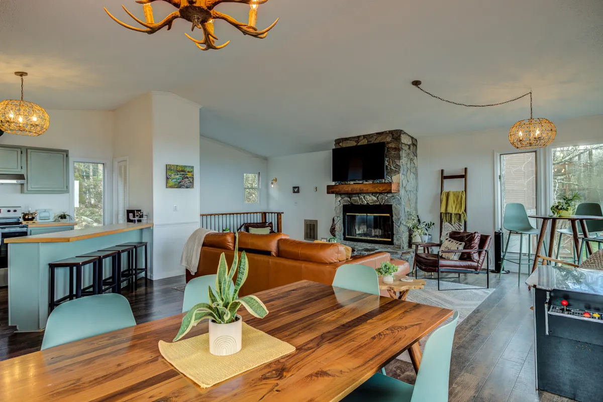 This view captures a broad interior shot with a dining table, leather sofa, fireplace, and part of the kitchen counter. The setting conveys warmth, togetherness, and the space’s flexibility for family meals or relaxed hangouts.