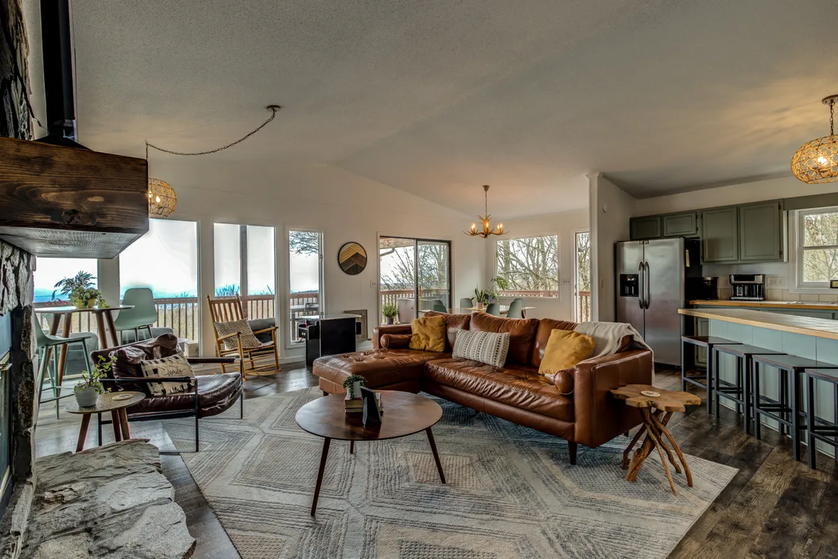 This image features a spacious interior living area with a leather sectional sofa, large windows framing stunning mountain views, a stone fireplace, and a round coffee table. The open layout hints at adjacent communal spaces, with abundant natural light and a cozy, inviting vibe.