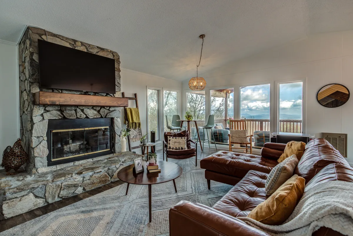 A welcoming living room with a stone fireplace, leather sofa, large windows framing the outdoors, and a spacious dining area. The scene suggests a spot where families can gather for games, relaxation, and to enjoy sunset views together.