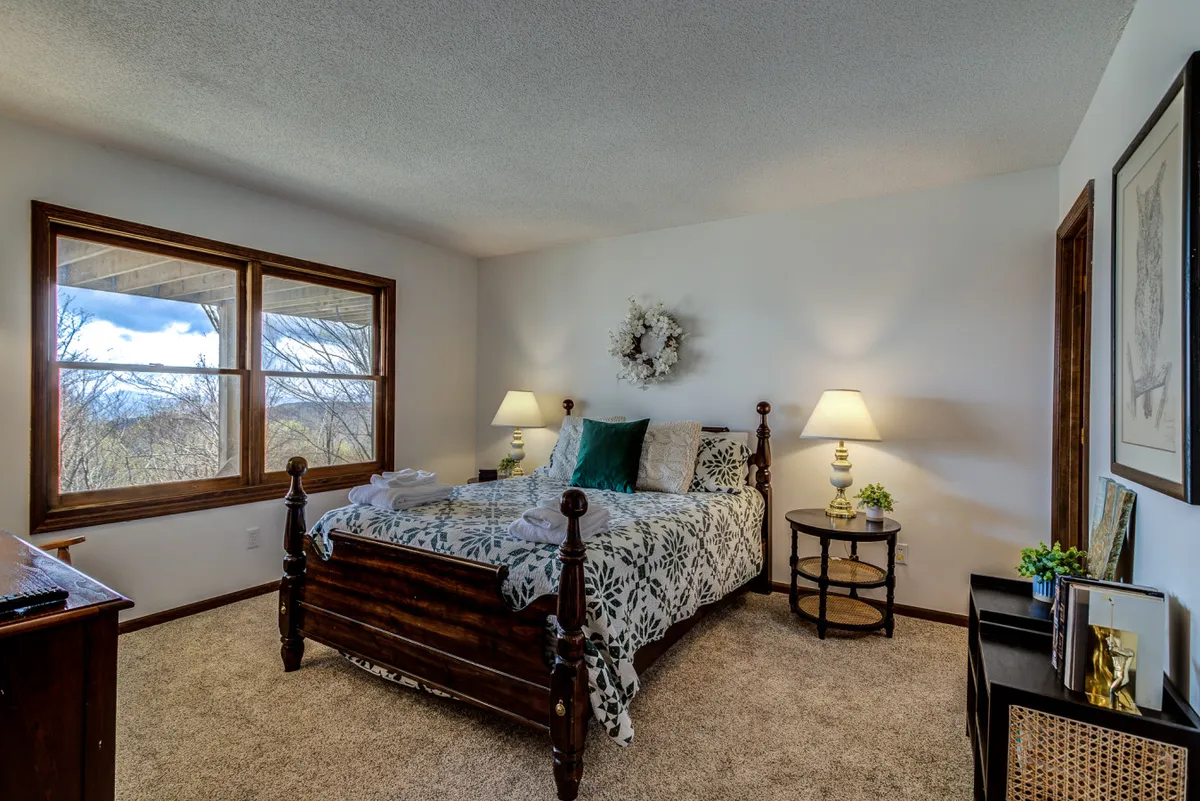A bright and inviting bedroom with a bed dressed in floral bedding, large windows offering abundant natural light, a decorative wreath above the bed, and a cozy nightstand with a lamp. The room exudes warmth and provides a sense of peaceful retreat.