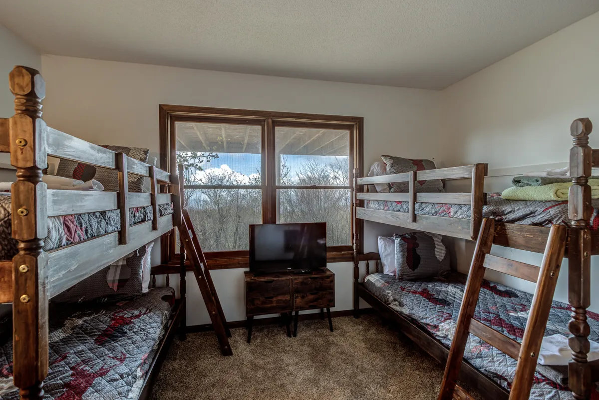 A playful bunk room with two twin-over-twin bunk beds, large windows letting in natural light, a wall-mounted television, and decorative pillows. The space is designed for both fun and relaxation, accommodating multiple guests or children.