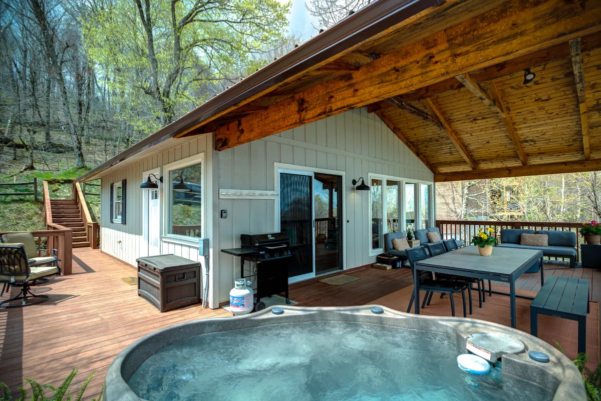 This exterior image highlights a large deck area featuring a bubbling hot tub, an outdoor dining table, lounge chairs, and a grill. The deck overlooks a natural setting, suggesting direct access to trails and ample space for relaxation, socializing, and dining in the fresh mountain air.