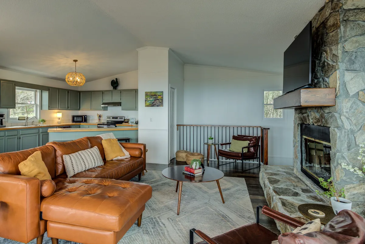 The image presents a cozy living area with a leather sectional sofa, a stone fireplace as the centerpiece, a wooden coffee table, and a view into the kitchen area. The seating is ample and plush, arranged for socializing or relaxing by the fire.