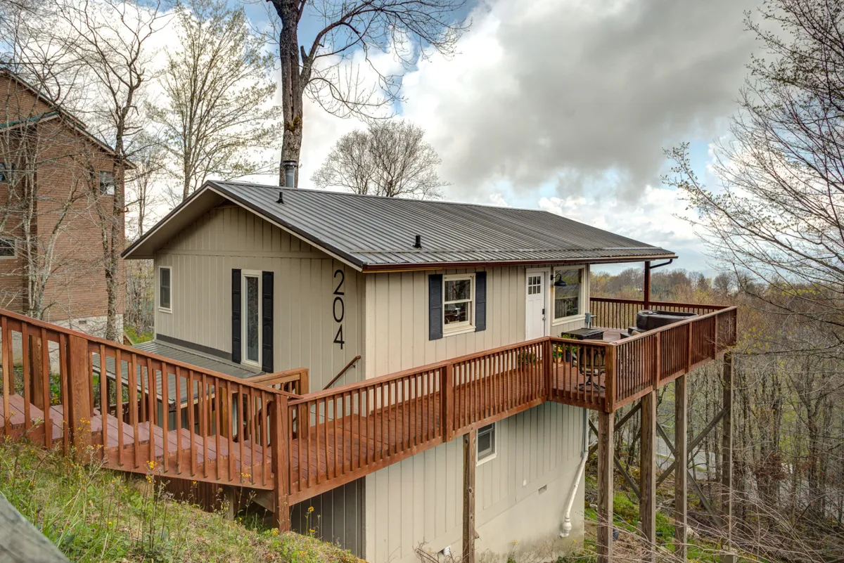 This exterior shot captures the raised deck and facade of the house nestled among lush trees. The setting is tranquil, with outdoor furniture arranged for meals and lounging, perfect for morning coffee or evening celebrations.
