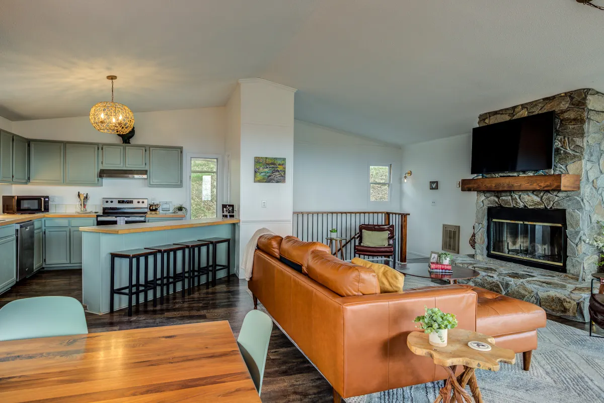 This image presents a warm, inviting living area featuring a leather sofa in front of a stone fireplace and a visible kitchen island in the background. The scene hints at a cozy retreat after a day outdoors, with room for relaxation and togetherness.
