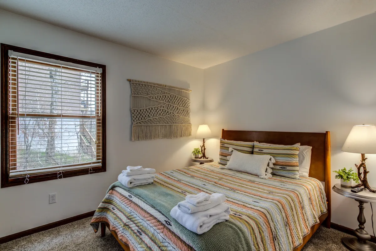 A cozy bedroom featuring a bed with colorful striped bedding, a decorative wall hanging above the headboard, and warm table lamps on each side. The overall ambiance is welcoming and tranquil, with soft lighting and comfortable textures inviting relaxation.