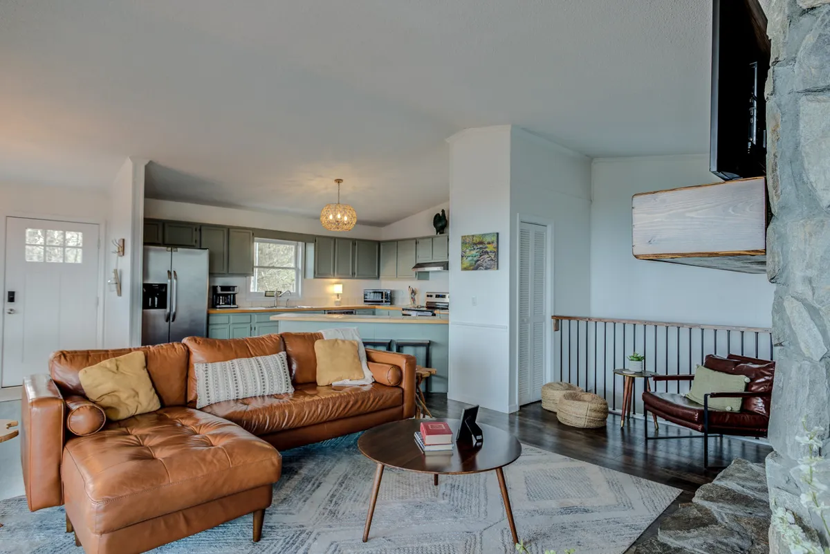 The image shows the heart of the home: a brown leather sectional sofa, round coffee table, decorative pillows, and a stone fireplace. The view extends into the open kitchen area, illustrating the seamless flow between the living and cooking spaces.
